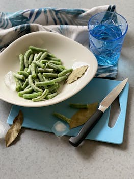 Frozen green beans in a bowl with knife and water glass on a cutting board.
