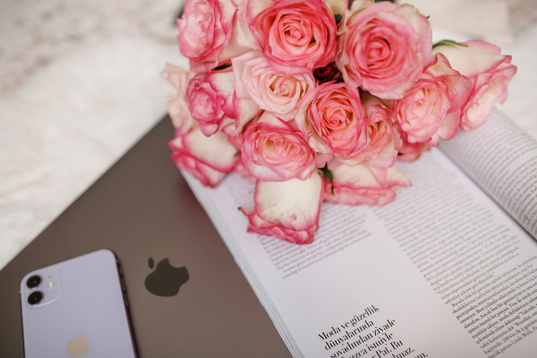 Aesthetic flat lay featuring a bouquet of pink roses, a laptop, and a smartphone on a magazine.