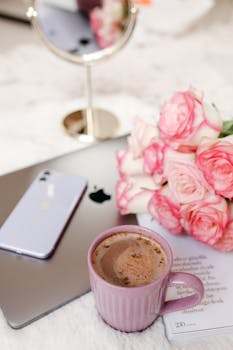 A delightful arrangement of a ceramic mug with cocoa, pink roses, a smartphone, and a laptop on a soft surface.