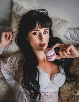 A beautiful woman in white lingerie holding a pink macaron, lounging luxuriously.