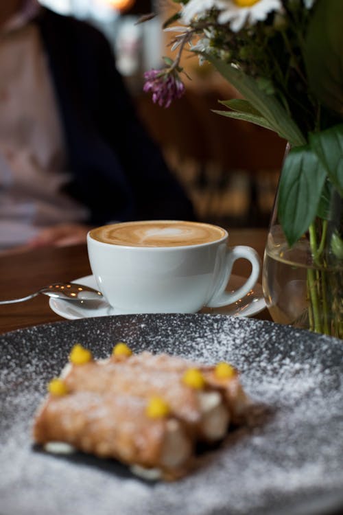 Free Cup of fresh latte placed near plate with cannoli pastry and vase with flowers during breakfast in cafe Stock Photo