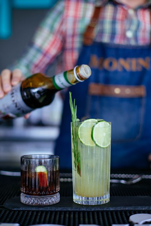 Free Unrecognizable bartender pouring gin from bottle into glass with slices of fresh lime and rosemary served near glass filled with tasty negroni cocktail in daylight Stock Photo