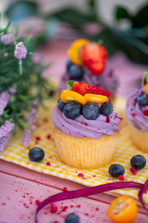 Cupcakes with purple cream decorated with fresh berries placed on yellow napkin near plant against blurred background