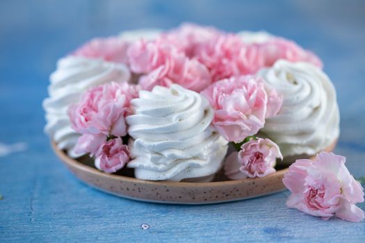 From above of delicious white marshmallows zephyr and buds of pink flowers placed on plate on blue table in room against blurred background