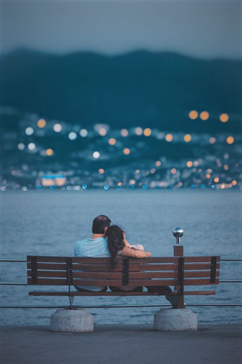 Free Couple embracing on a bench by a lake with city lights in the background. Stock Photo