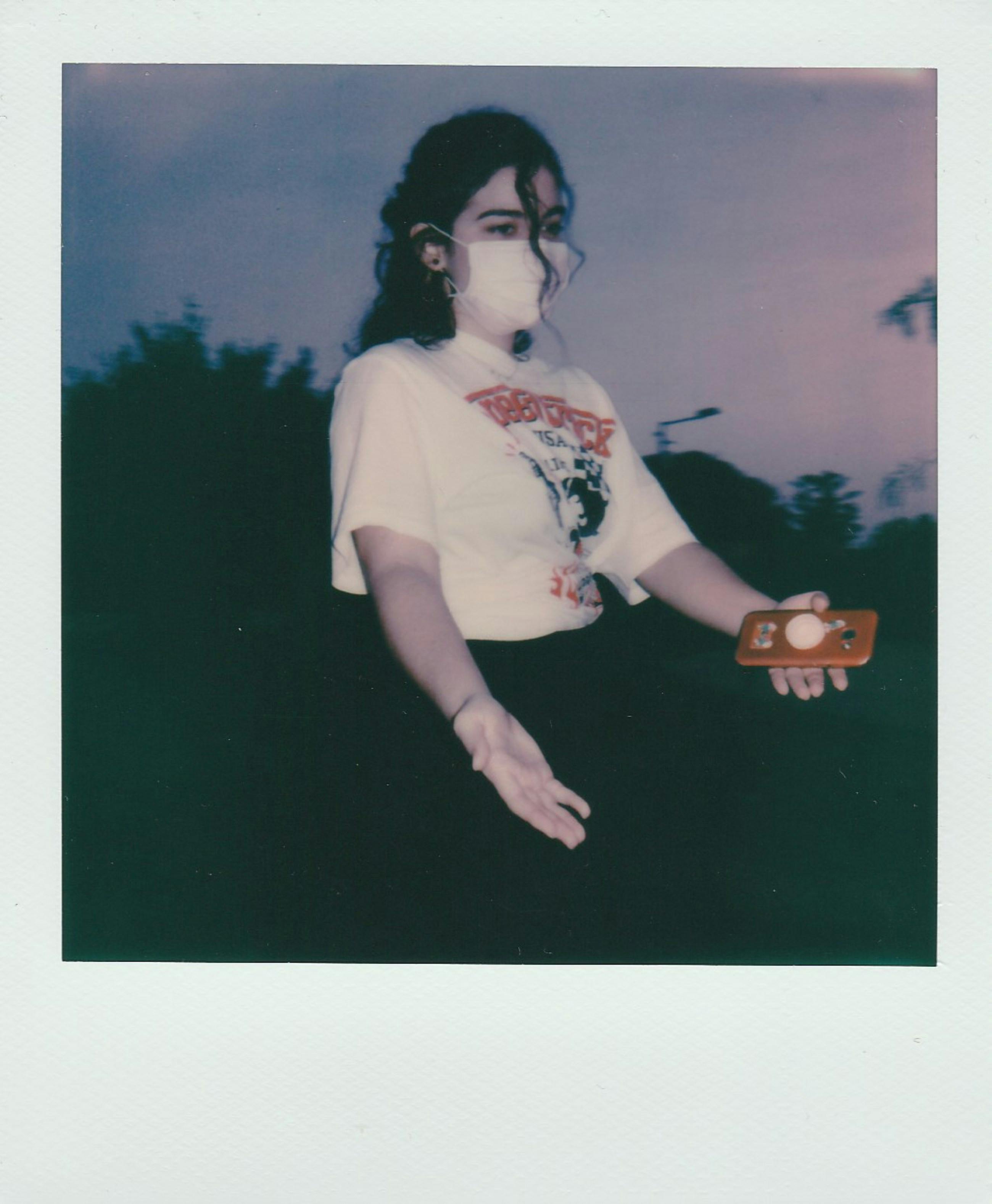 A young woman wearing a face mask outdoors holding a smartphone, set against a twilight sky.