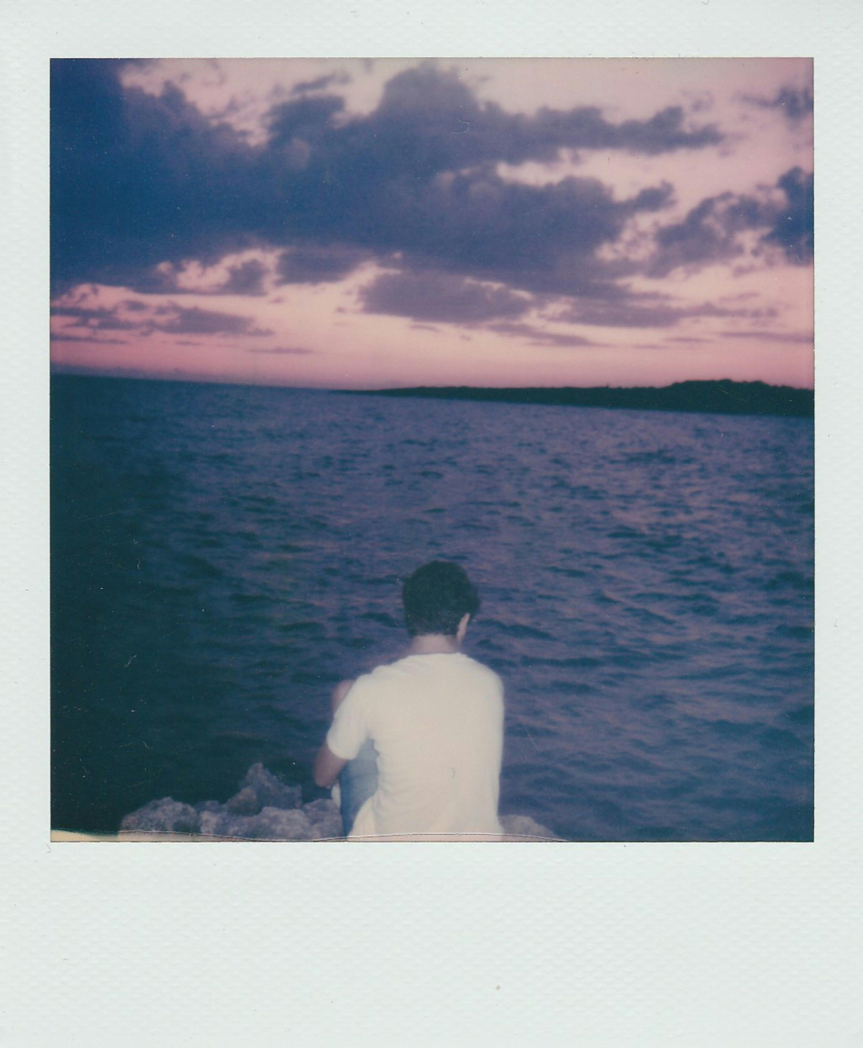 A serene view of a man sitting by the sea at sunset with a vibrant cloudy sky in Taranto.