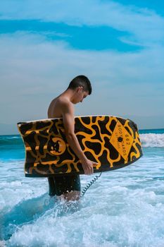 A young man stands in the surf holding a colorful bodyboard under a bright blue sky.