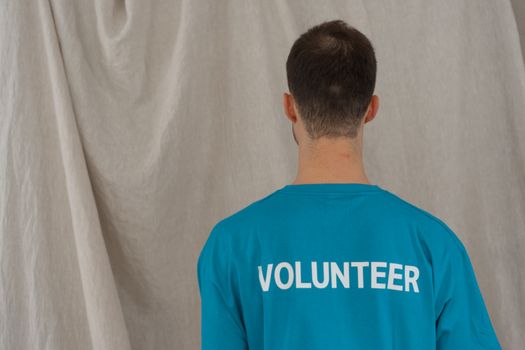 A man in a blue volunteer shirt seen from behind, indoors.