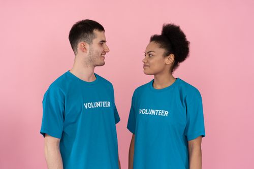 Free Two diverse volunteers smiling at each other, wearing blue shirts with 'VOLUNTEER' text against a pink background. Stock Photo