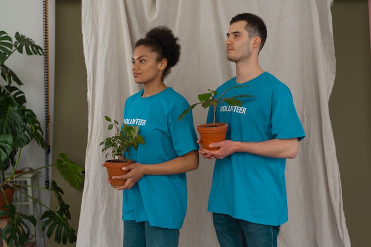 A Man And Woman In Blue Crew Neck T-shirts Holding Plants In Brown Pots