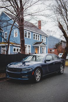 Modern expensive automobile parked on asphalt road near fence of cottage with blue walls located in residential district in town in autumn time in daylight