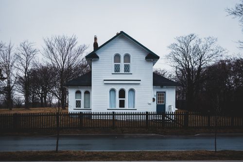 Free White house with ornamental windows and leafless trees behind wooden fence located near wet asphalt road in overcast day Stock Photo