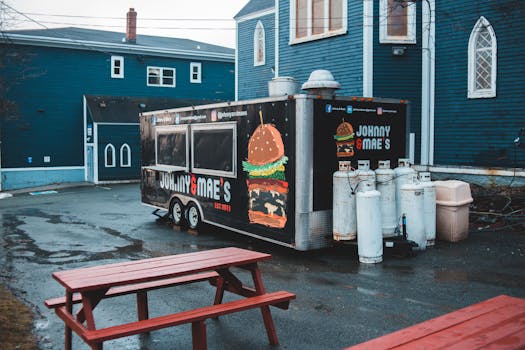 From above of food truck placed on wet asphalt area with wooden tables and benches in front of blue buildings