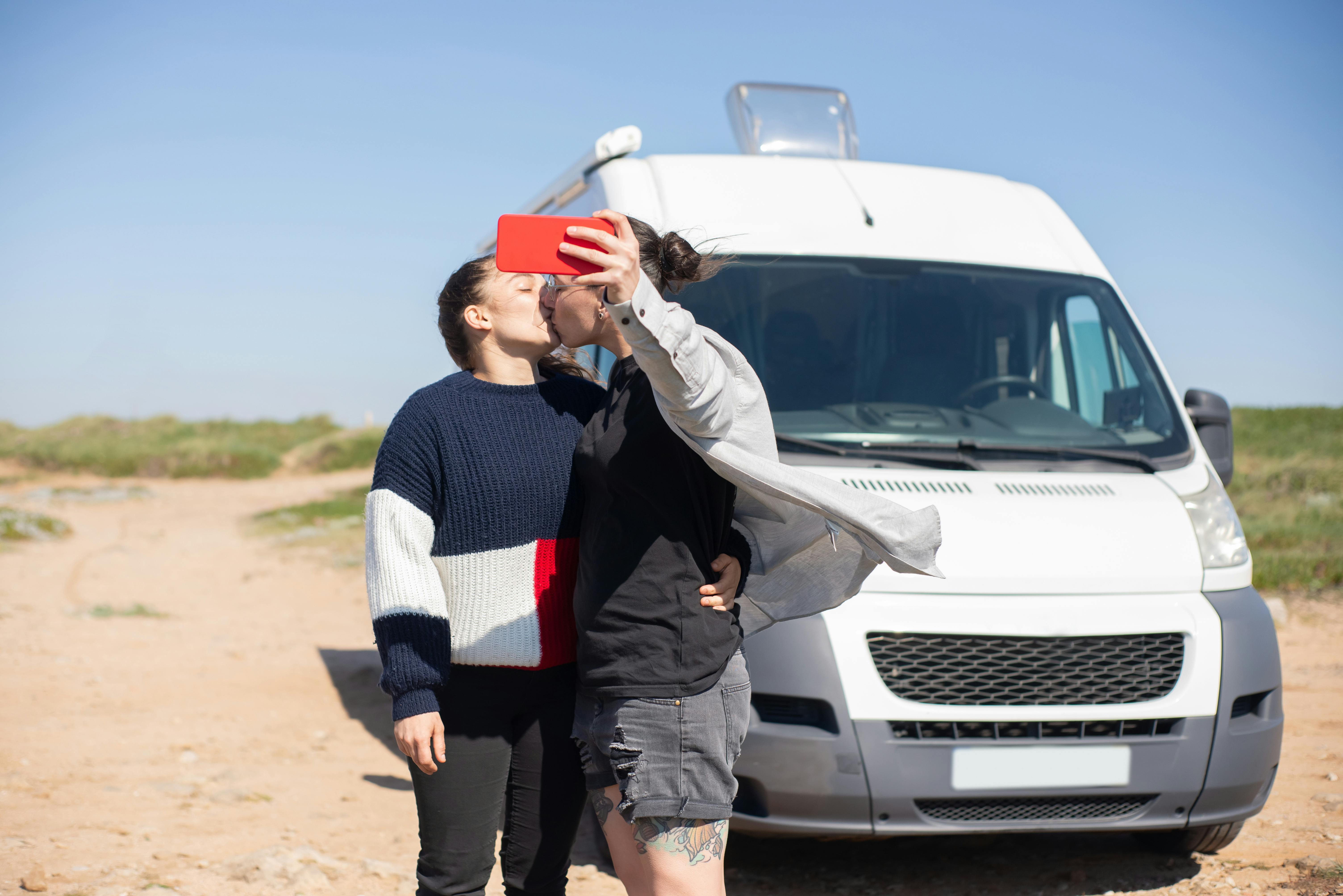 A couple shares a kiss while taking a selfie in front of their camper van, enjoying a sunny day outdoors.