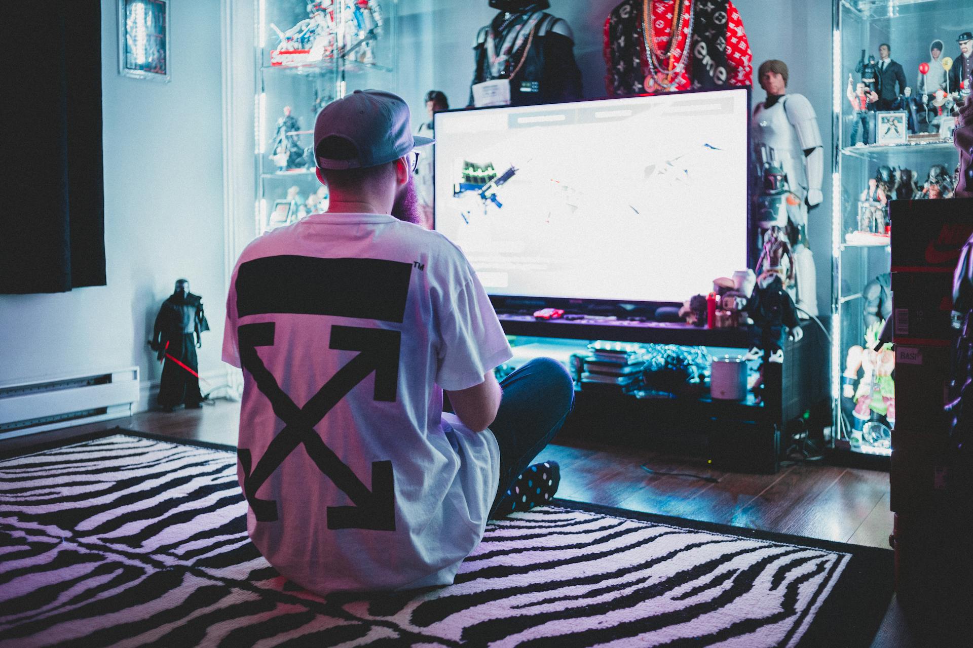 Back view of anonymous geek male in casual clothes and cap sitting on carpet and playing console video game in living room full of toys and stuff from comic