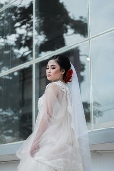 Elegant Asian bride in a white wedding gown with red rose accent posing outdoors in natural light.