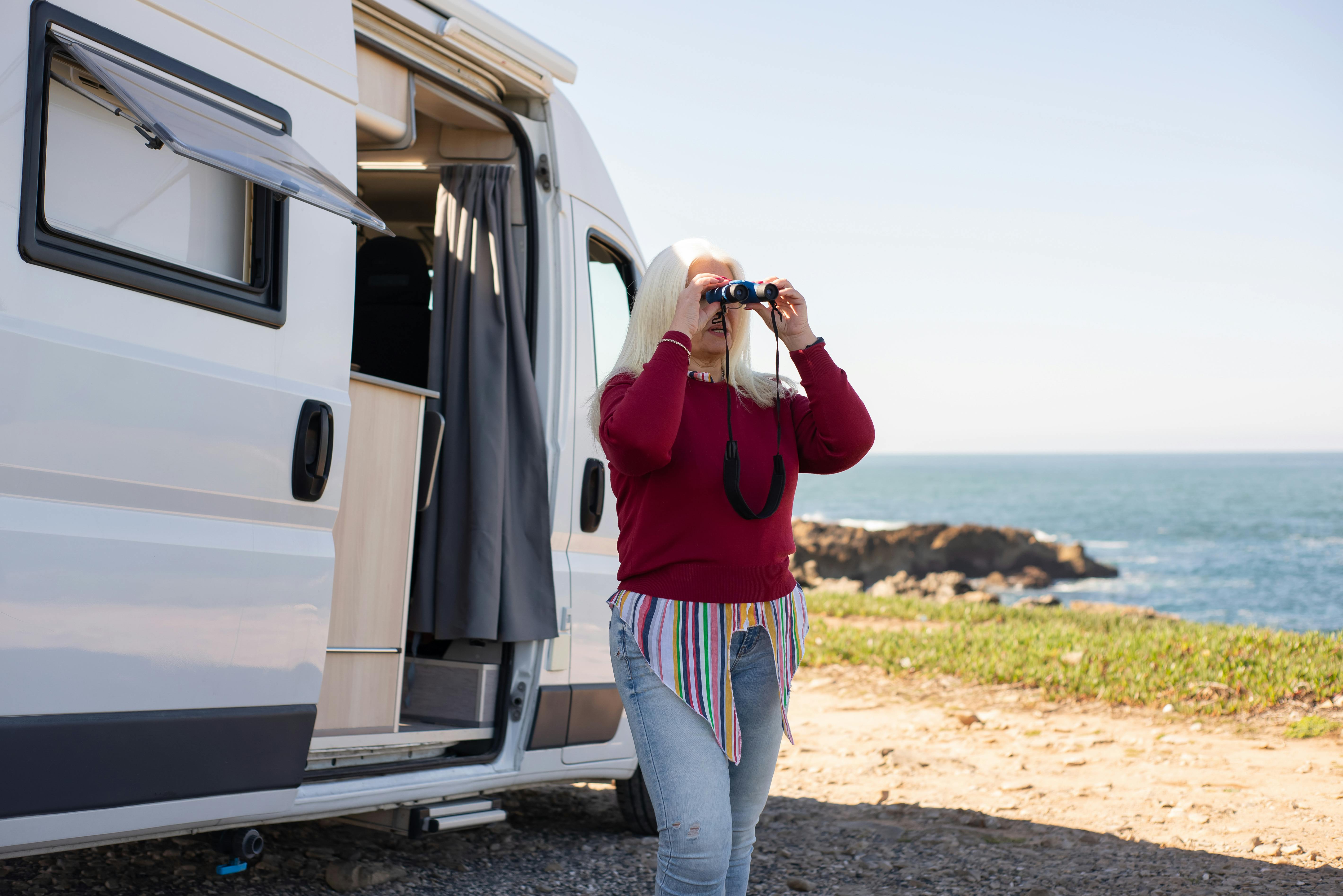 Woman enjoying a scenic view by the sea with binoculars near a camper van in Portugal.