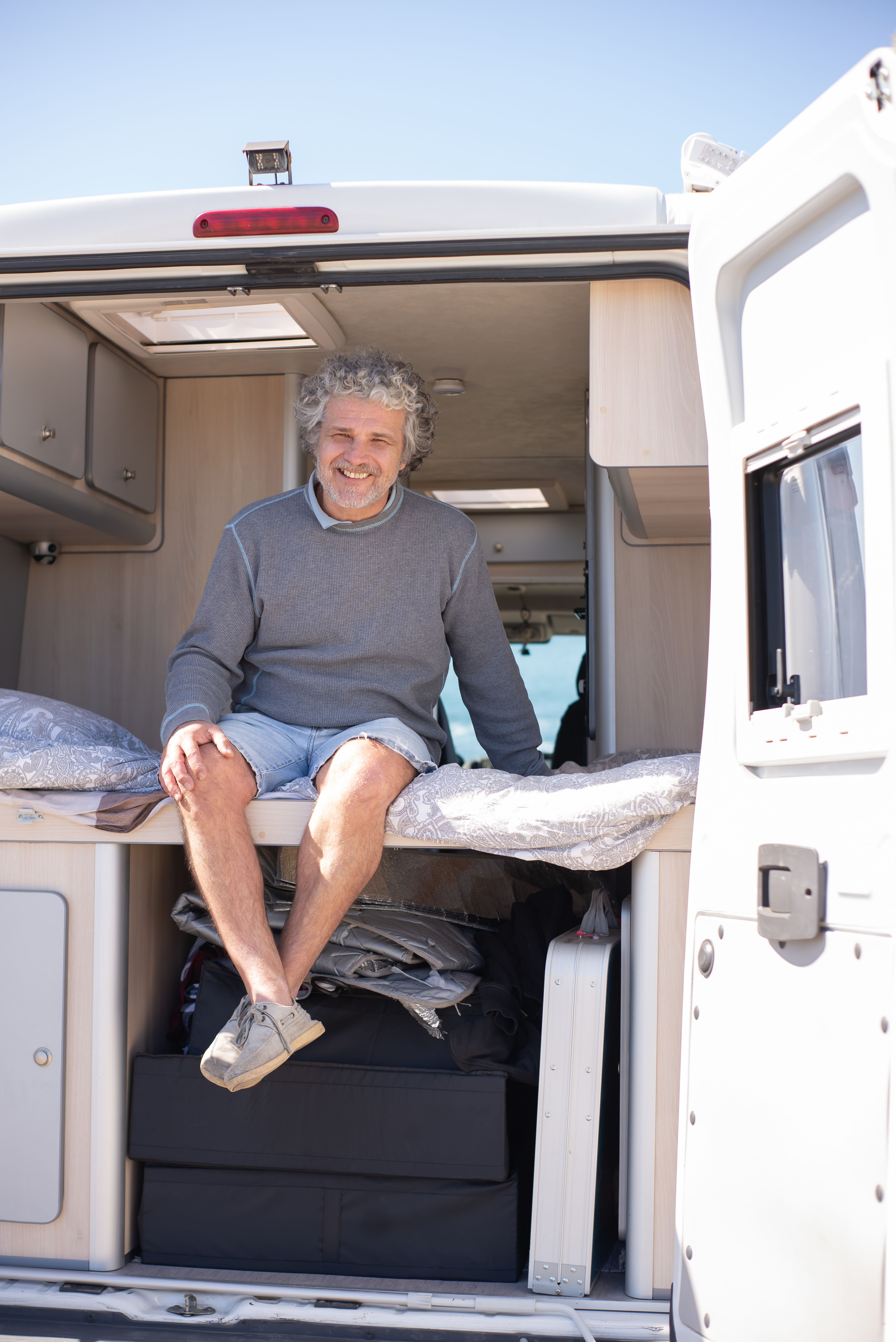 Senior man with gray hair sits smiling in a camper van on a sunny day in Portugal.
