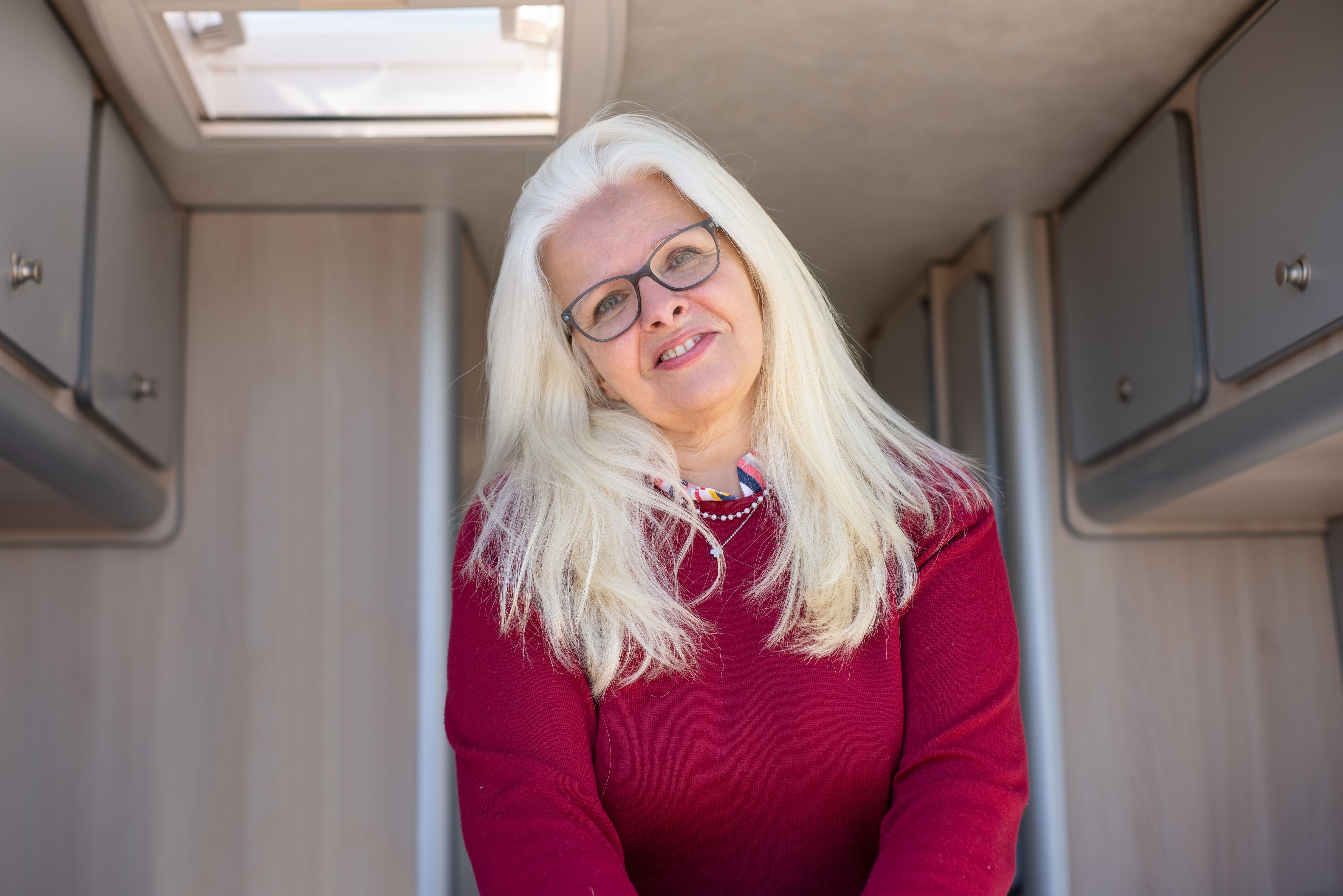A cheerful woman with glasses and white hair smiling warmly inside a cozy camper van.