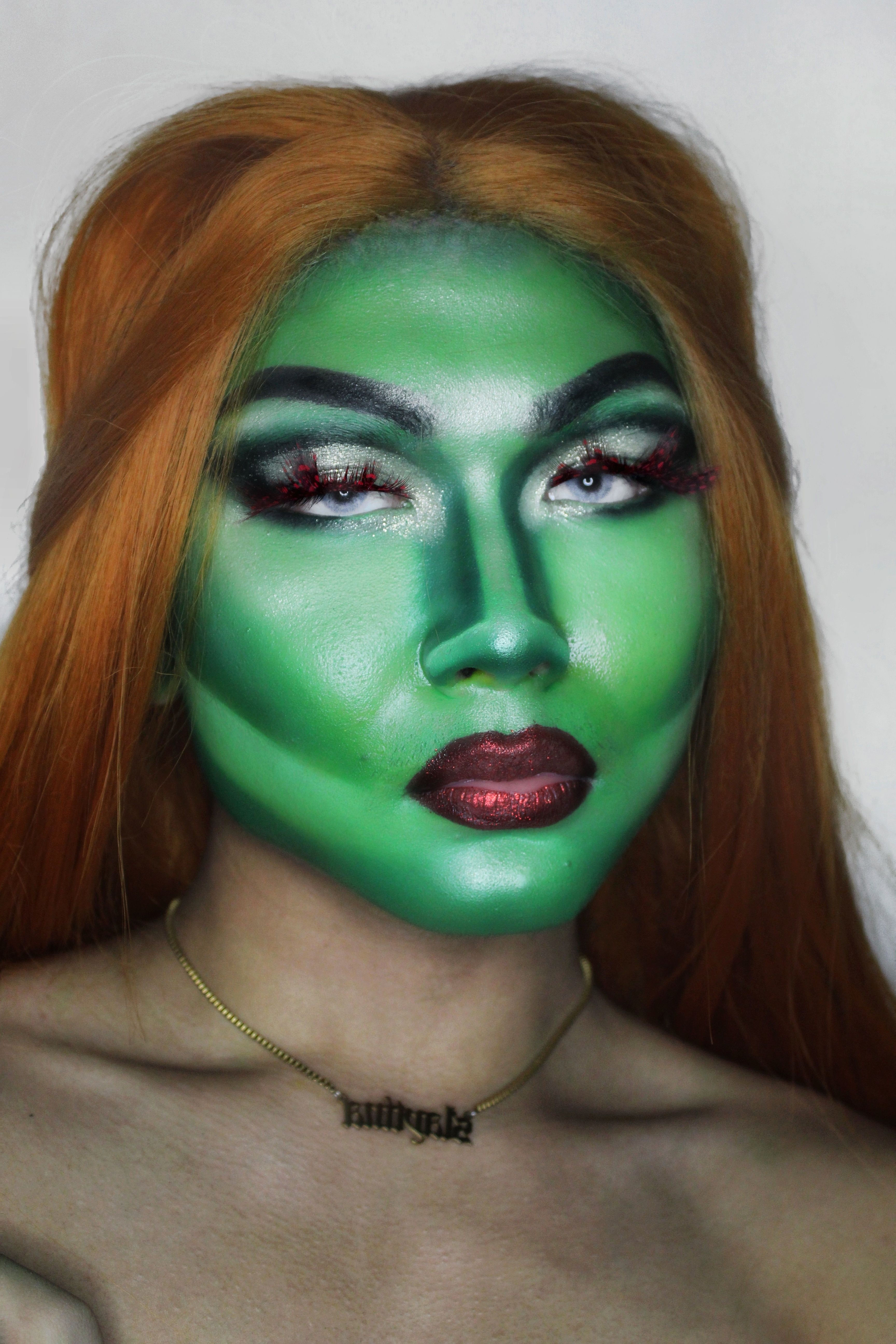 Striking portrait of a woman with artistic green facial paint and long orange hair, against a white background.