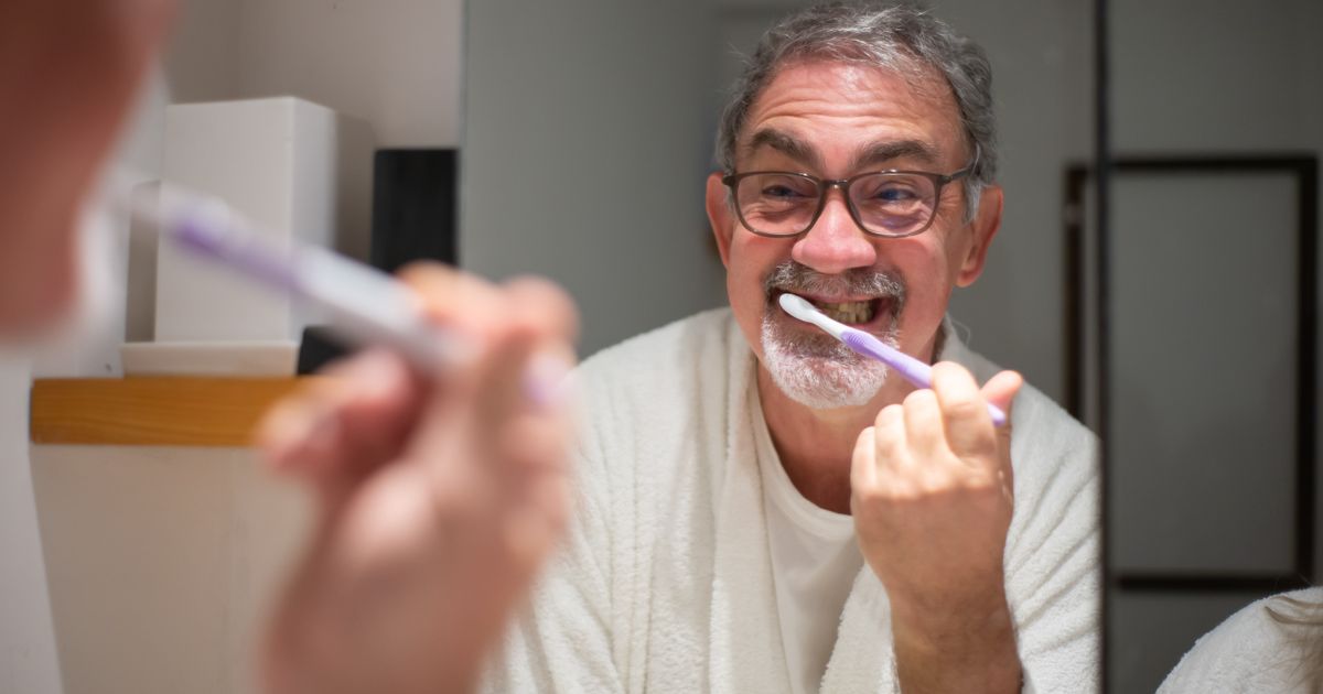 An older man smiling while brushing his teeth in front of a bathroom mirror
