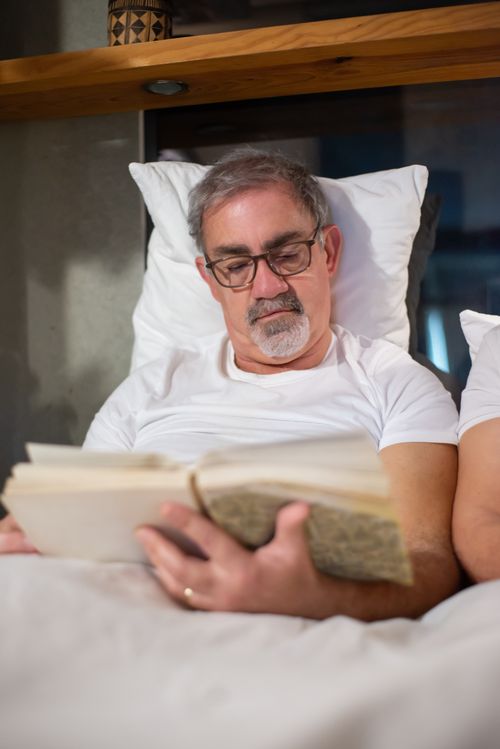 Free Elderly man reading a book in bed, enjoying leisure time with glasses on. Stock Photo