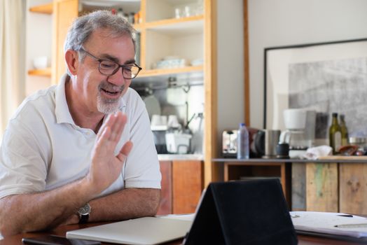 Smiling elderly man using digital tablet for video chat in cozy home kitchen setting.