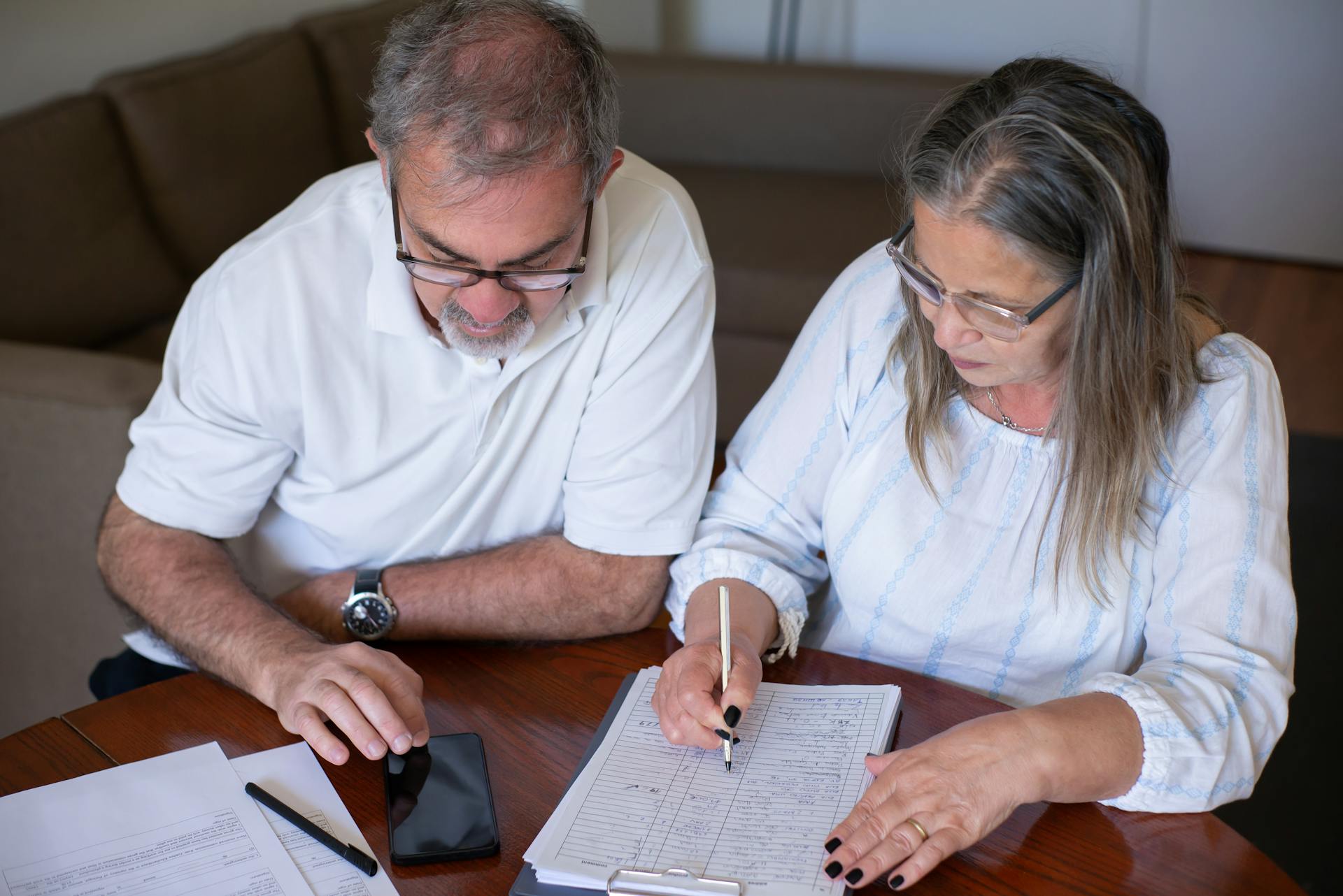 Elderly couple reviewing documents, using smartphone for online banking at home.