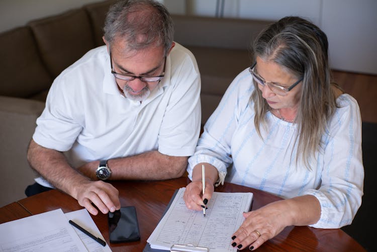 Elderly Couple Sitting At The Table With Documents And Using A Smartphone