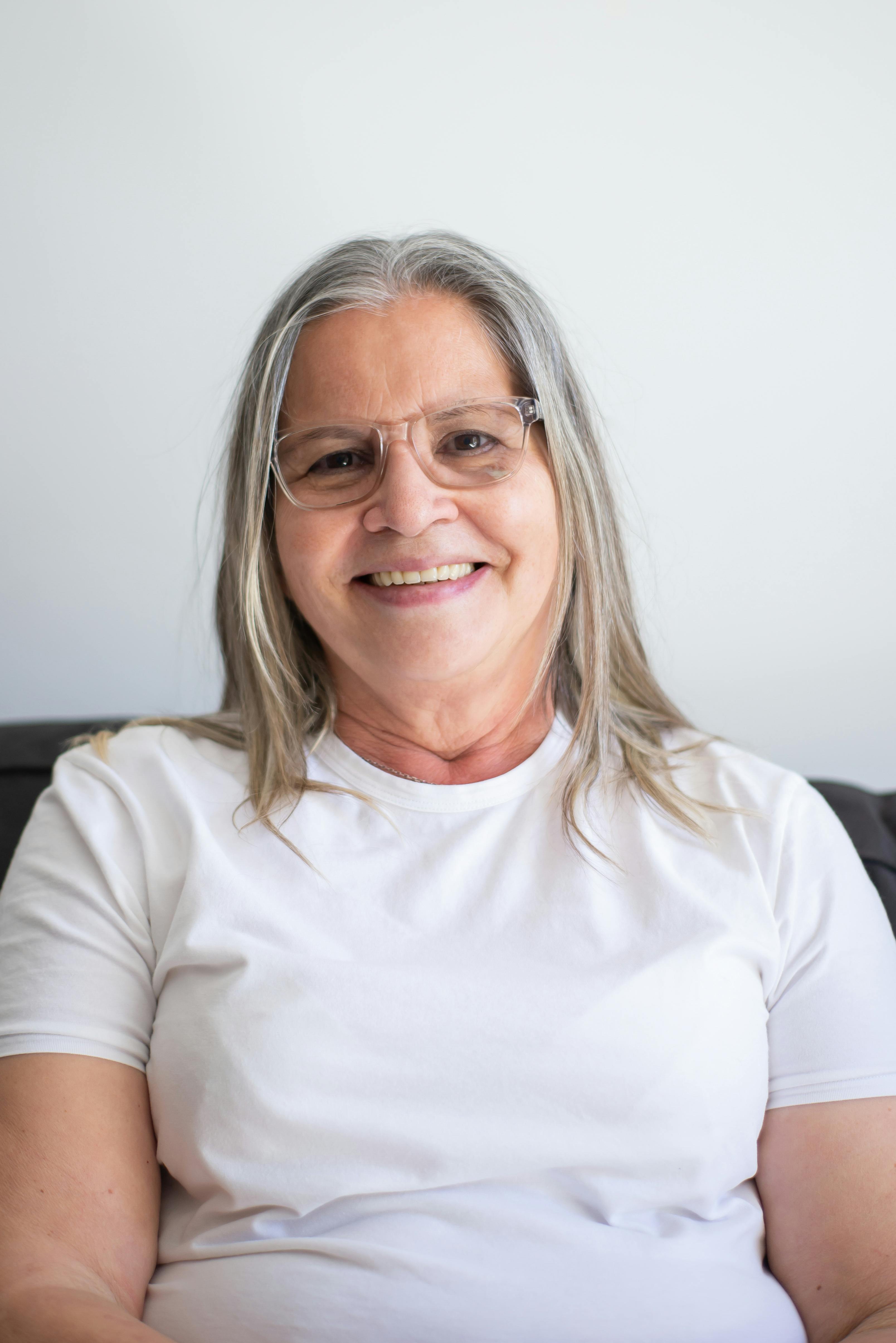 Portrait of a smiling elderly woman wearing eyeglasses and a white t-shirt indoors.