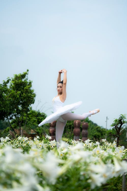 Free Professional flexible ethnic female artist in tutu dress doing ballet movement with raised arms in blooming meadow Stock Photo