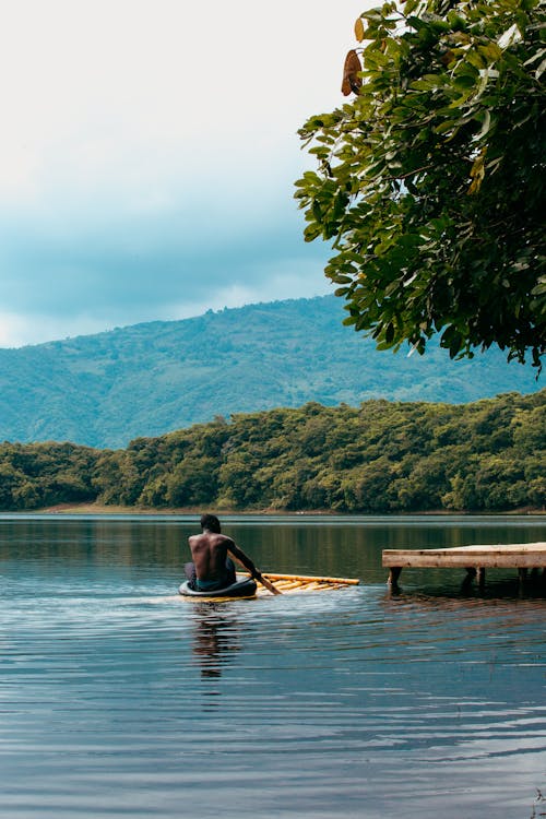 Back view of anonymous ethnic male on bamboo raft with oar sailing on rippled lake against green mount