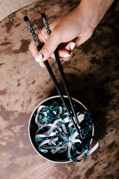Hand using chopsticks to hold decorative ribbon in a bowl, viewed from above.