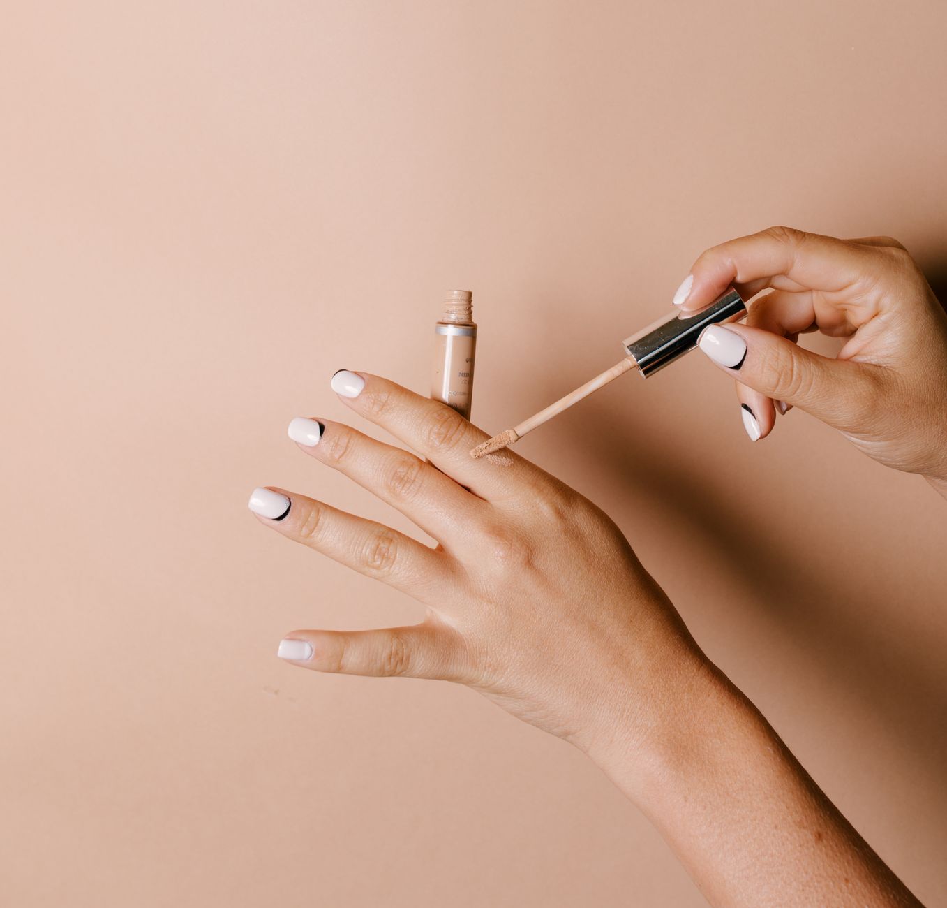 Elegantly manicured hands applying liquid concealer on skin against a neutral background.