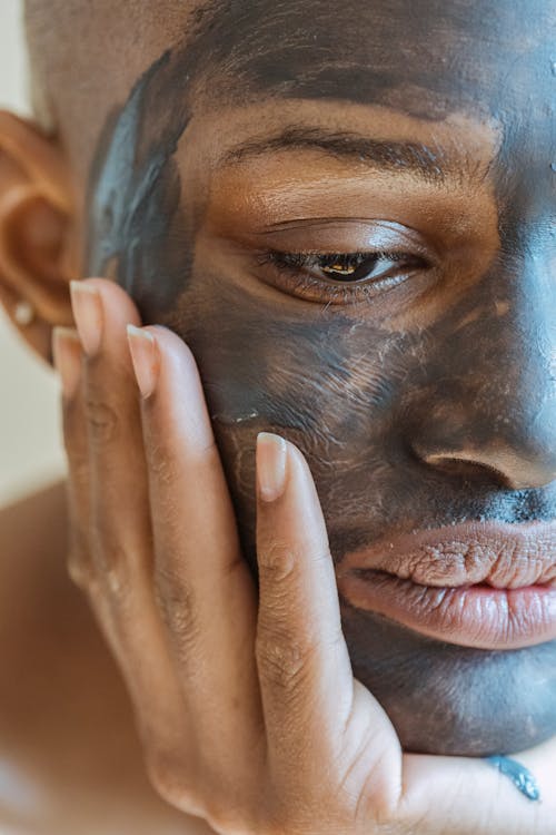 Crop sad African American female touching face while doing skin care clay mask and looking away with bored glance