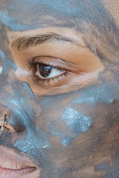 Closeup of crop African American female with brown eyes taking care of skin with mud mask