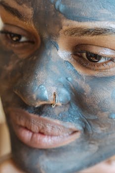 Crop Black female with pierced nose and mud mask on face looking away during spa procedures