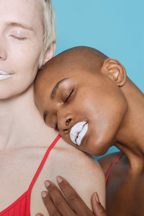 Free African American woman with closed eyes and white lipstick leaning head on bare shoulder of crop model on blue background Stock Photo