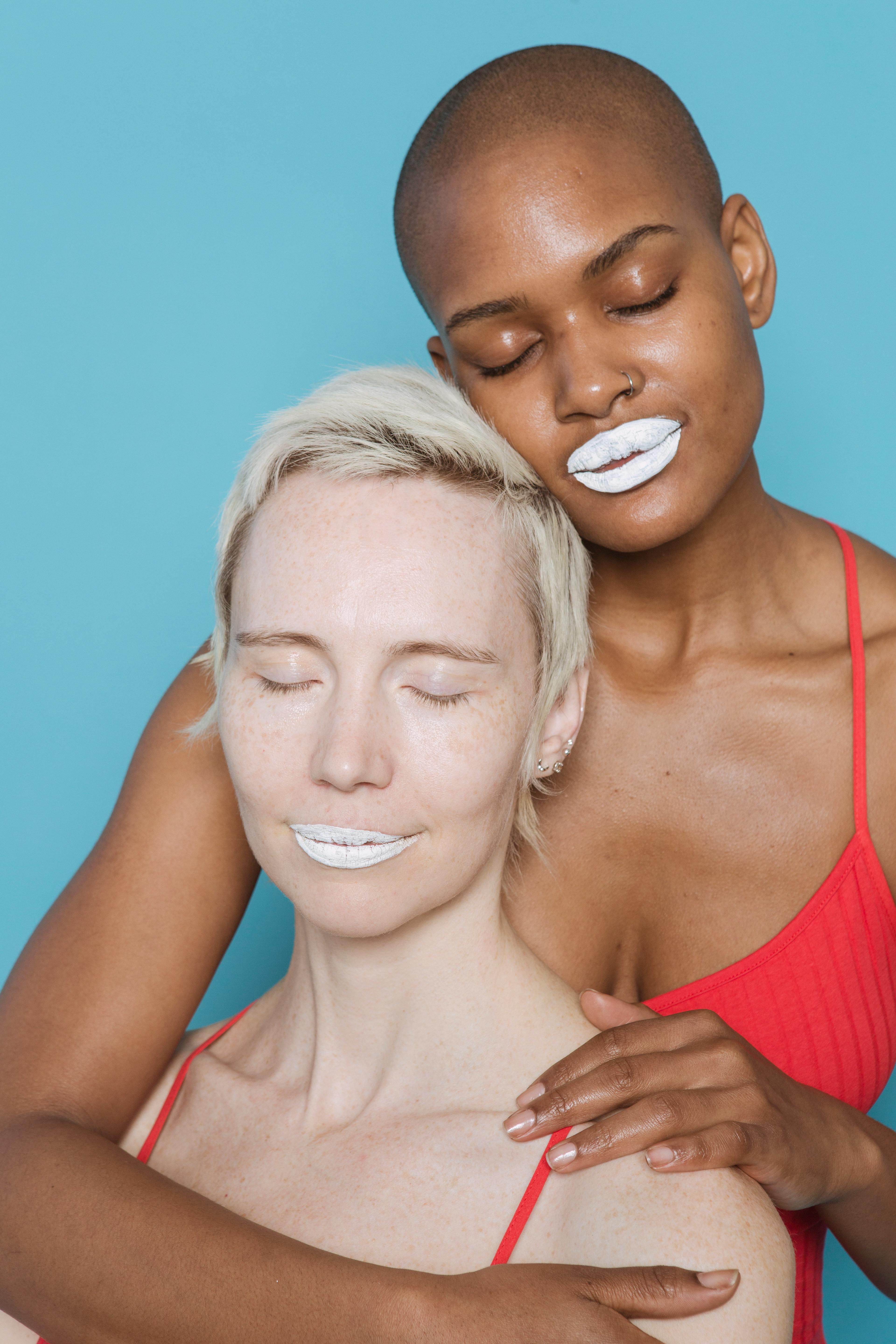 Dreamy multiracial adult women in red bodysuits with white lips and closed eyes cuddling on blue background in bright studio