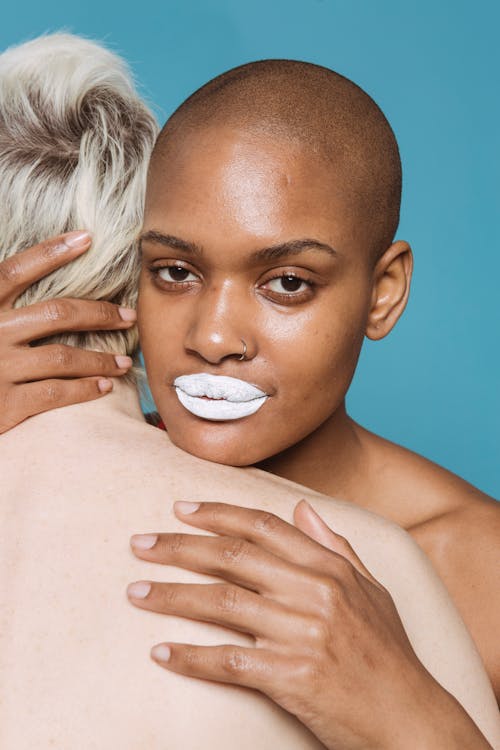 Multiethnic adult women with white lipstick and bare shoulders embracing each other in light studio on blue background