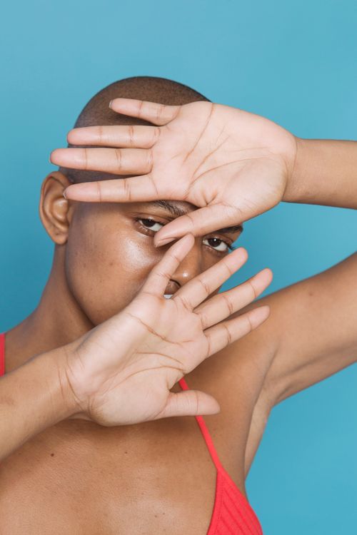 Free Bald black female in bra looking at camera while covering face with hands on blue background in light studio Stock Photo
