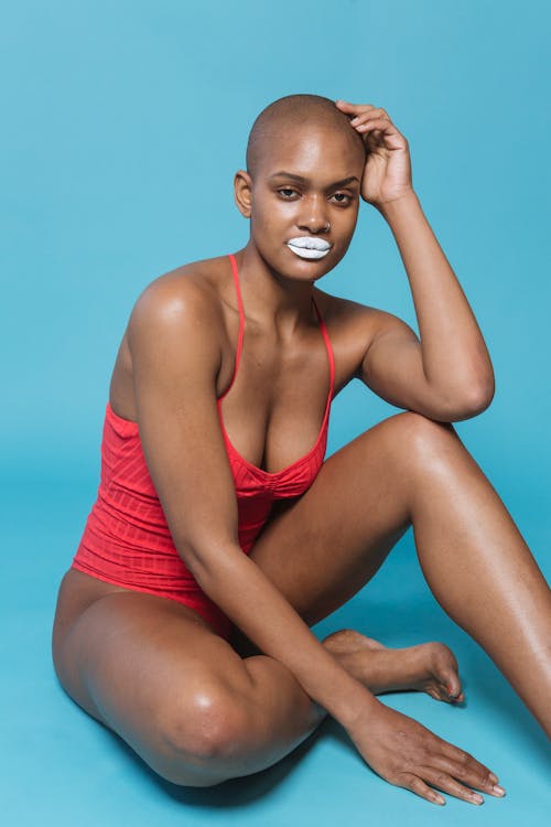 Serious black lady in swimsuit with white lips and bald head sitting on blue background while looking at camera with hand on head in bright studio