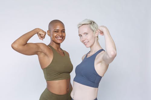 Cheerful diverse fit females in sportswear pumping biceps muscle and looking at camera while standing against white background