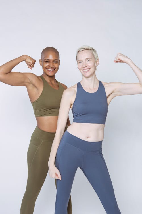 Glad slim diverse ladies in sportswear standing with flexed arms and looking at camera on white background