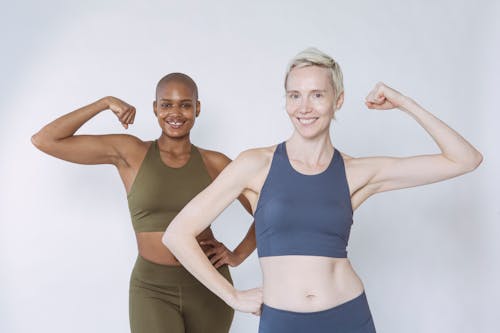 Smiling multiracial women in workout clothes standing against white background and flexing arms while looking at camera