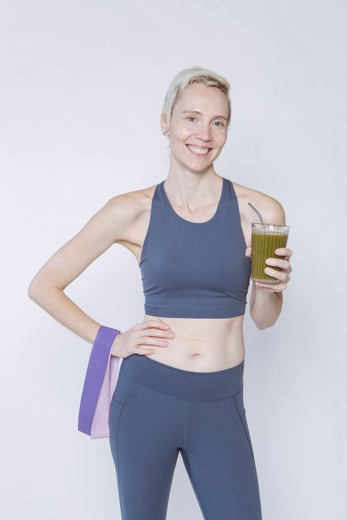 Free Positive female athlete with hand on waist standing with glass of vegetable smoothie and resistance band against white background Stock Photo