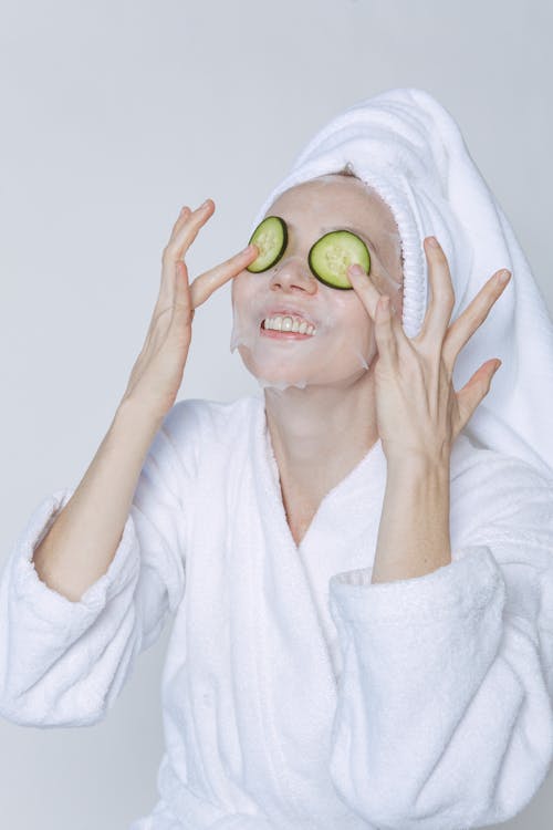 Young female in bathrobe and towel taking care of skin with cucumber and sheet mask against white background