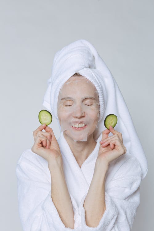 Positive female in white bathrobe and turban holding cucumber slices while moisturizing skin with sheet mask against white background