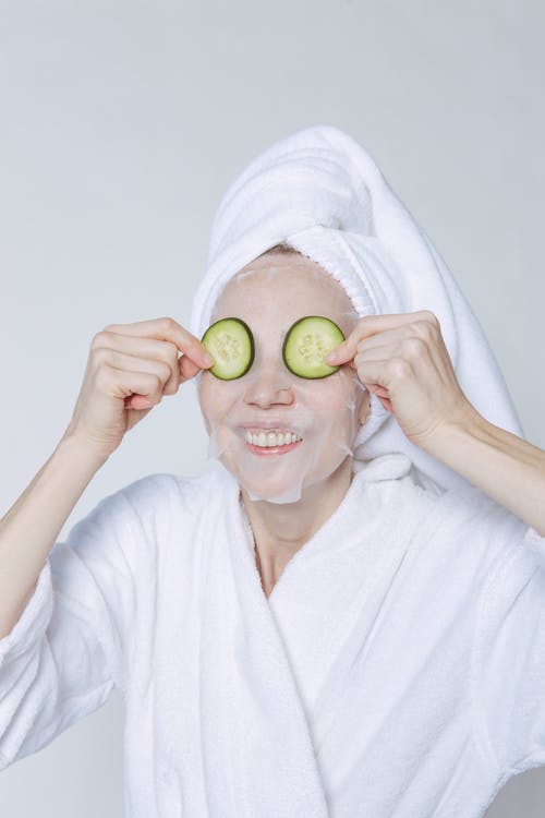 Positive female wearing white bathrobe and towel smiling happily while applying cucumbers on eyes for moisturizing skin against white background